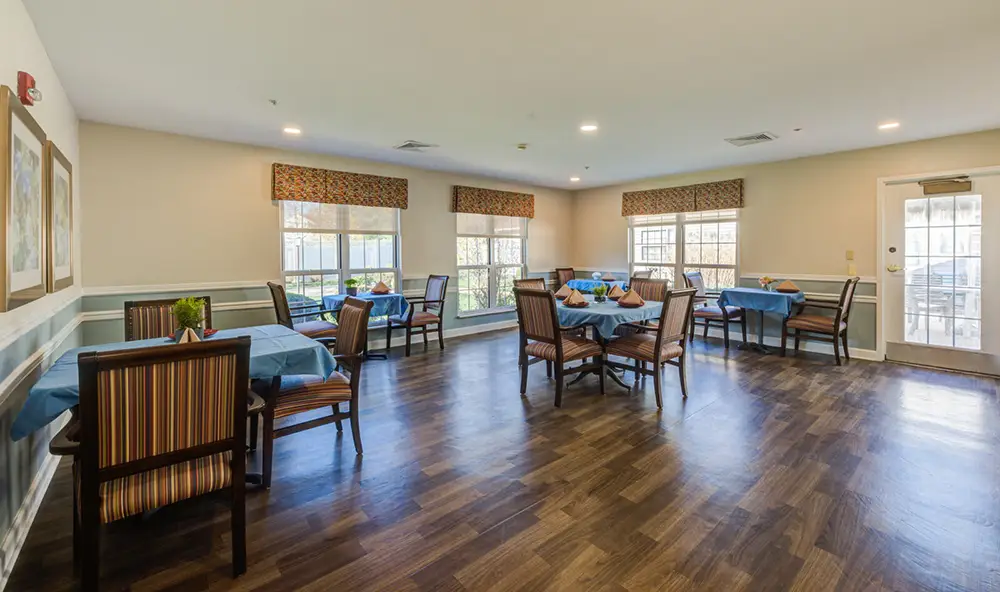 A dining room with several tables covered in blue tablecloths, wooden chairs, large windows, and wood flooring in a well-lit space.