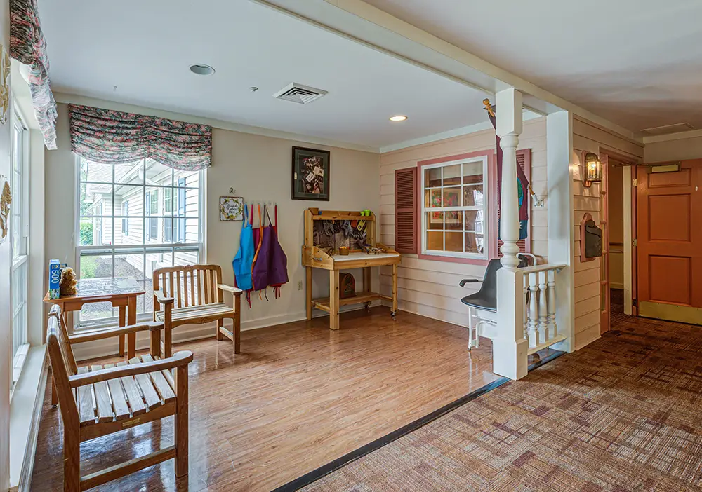 A well-lit room with wooden benches, aprons hanging on hooks, a craft table with art supplies, and a large window overlooking an outdoor area. A well-lit room with wooden benches, aprons hanging on hooks, a craft table with art supplies, and a large window overlooking an outdoor area.