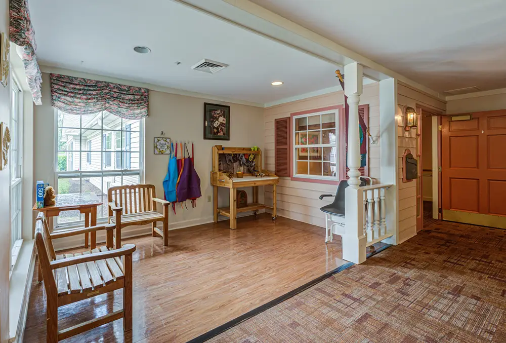 A cozy indoor space with wooden benches, an activity desk with aprons, a window, and a door, all on a mix of wood and carpet flooring.