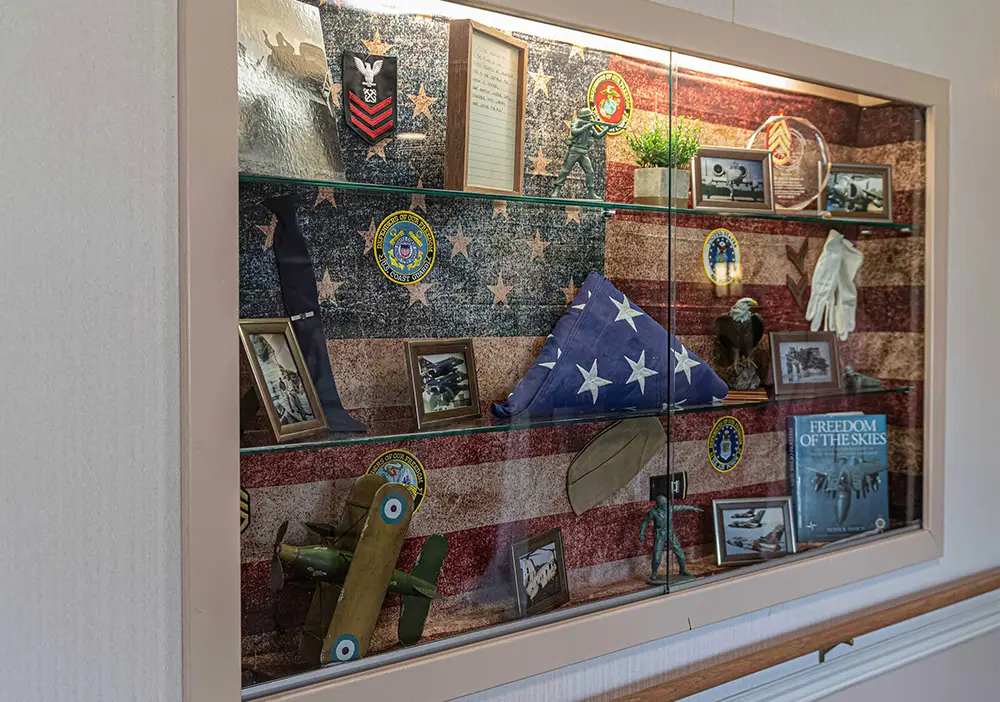 A display case contains military memorabilia, including a folded American flag, framed photos, model airplanes, patches, gloves, and plaques, set against an American flag background. A display case contains military memorabilia, including a folded American flag, framed photos, model airplanes, patches, gloves, and plaques, set against an American flag background.