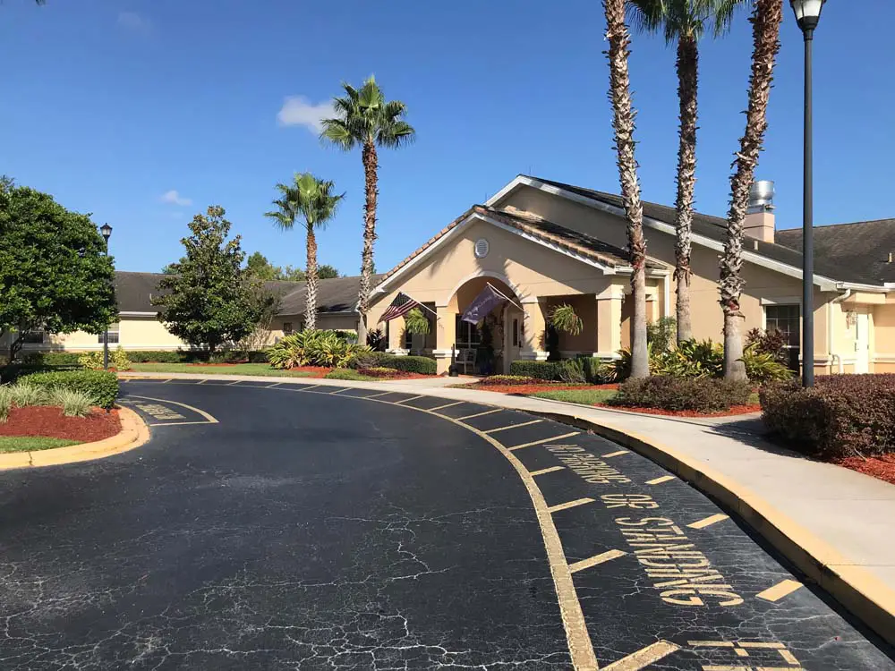 A single-story Arden Courts building in Winter Springs features a circular driveway, palm trees, and manicured landscaping under a clear blue sky. “No Parking or Standing” is painted on the asphalt, reflecting its dedicated memory care environment.
