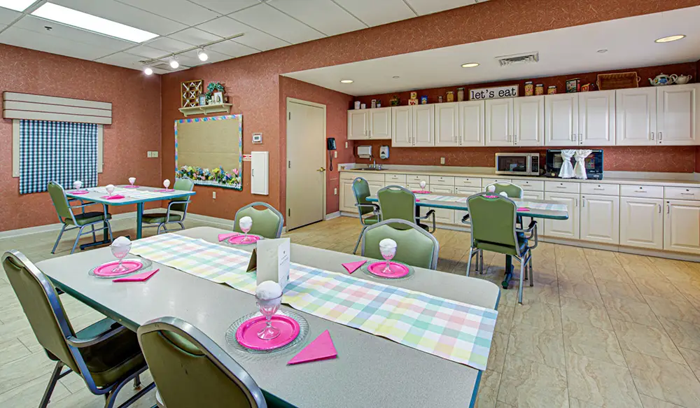 A dining area with green chairs, tables set with pink plates and napkins, white cabinets, and a bulletin board on a red wall.