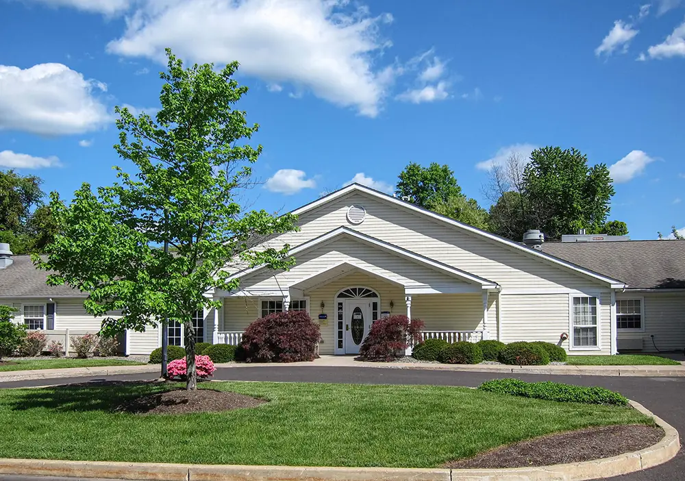 Single-story Arden Courts building with white siding and a central entrance, surrounded by landscaped bushes, a small tree, and a circular driveway—providing dedicated memory care in Yardley under a blue sky with scattered clouds.