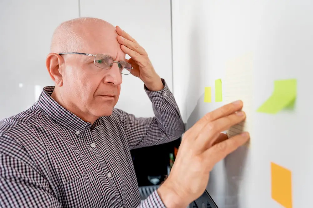 An older man wearing glasses looks at sticky notes on a wall, appearing thoughtful with one hand on his forehead and the other hand touching the notes. An older man wearing glasses looks at sticky notes on a wall, appearing thoughtful with one hand on his forehead and the other hand touching the notes.