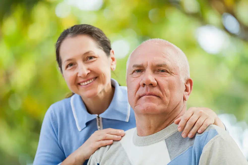 A woman smiles with her hands on the shoulders of an older man, who has a neutral expression, outdoors with greenery in the background at Arden Courts Memory Care. A woman smiles with her hands on the shoulders of an older man, who has a neutral expression, outdoors with greenery in the background at Arden Courts Memory Care.
