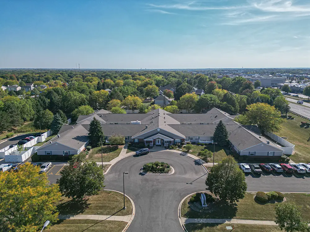 geneva 2 Aerial view of a large, single-story building complex with a circular driveway, surrounded by trees, parked cars, and suburban landscape under a clear blue sky.