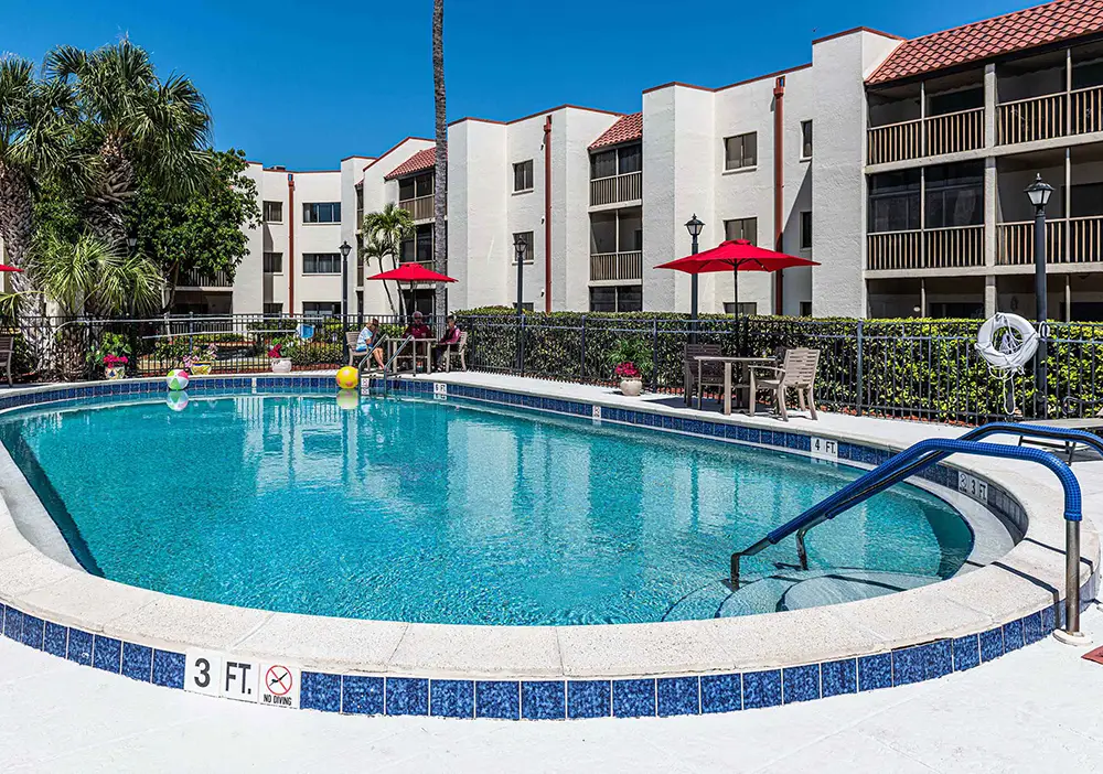 Enjoy the outdoor swimming pool at Lely Palms Senior Community, featuring clear water, a white deck with lounge chairs, red umbrellas, and three-story apartment buildings in the background.