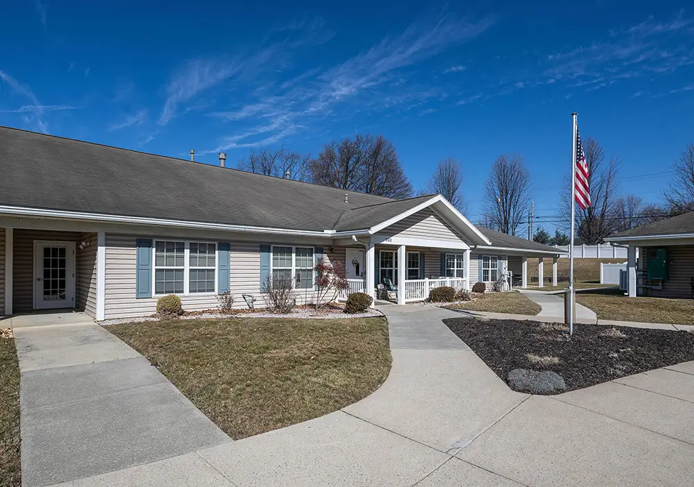 Single-story Linden Village building with light siding and multiple windows, surrounded by walkways, sparse landscaping, and an American flag on a pole, under a clear blue sky.