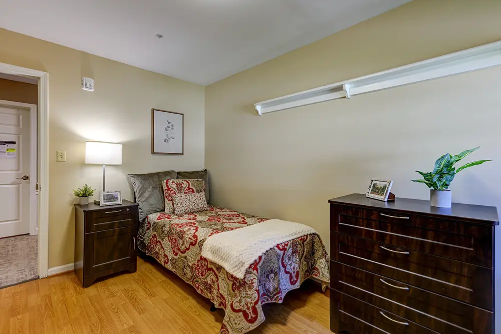 untitled-1Kensington-3 A neatly made single bed with patterned bedding is next to a nightstand and lamp in a simple bedroom with wood flooring and beige walls.