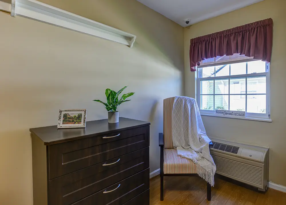untitled-1Kensington-4 A small room with a dark wood dresser, a framed photo, a potted plant, an armchair with a draped blanket, and a window with a maroon valance above an air conditioning unit.